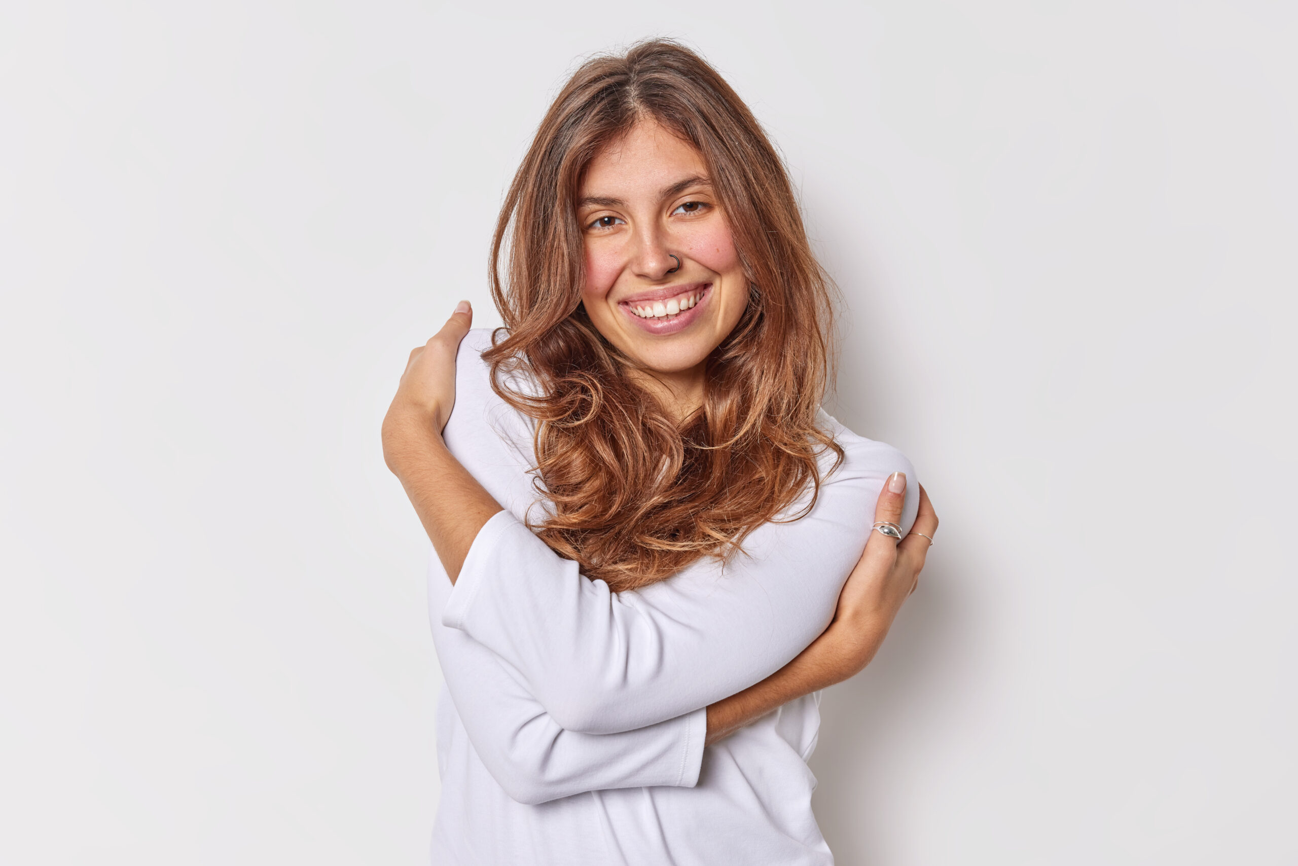 Tender optimistic long haired European woman embraces herself expresses self love and acceptance smiles happily wears casual jumper isolated over white background. Coziness tenderness concept